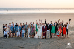 Joyeux groupe de mariage rassemblé sur le sable doré, avec l'océan bleu azur en toile de fond, capturant l'amour et la camaraderie partagés lors de cette journée mémorable.
